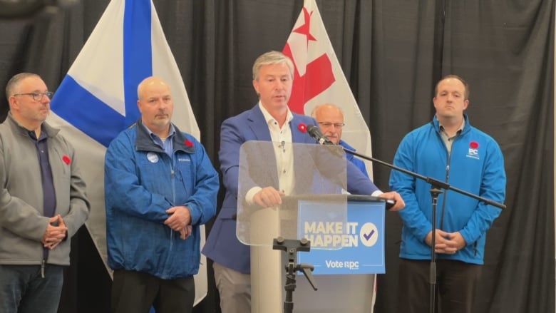 A group of men stand in front of Nova Scotia and Mi'kmaw flags while a man in blue blazer speaks at a podium.