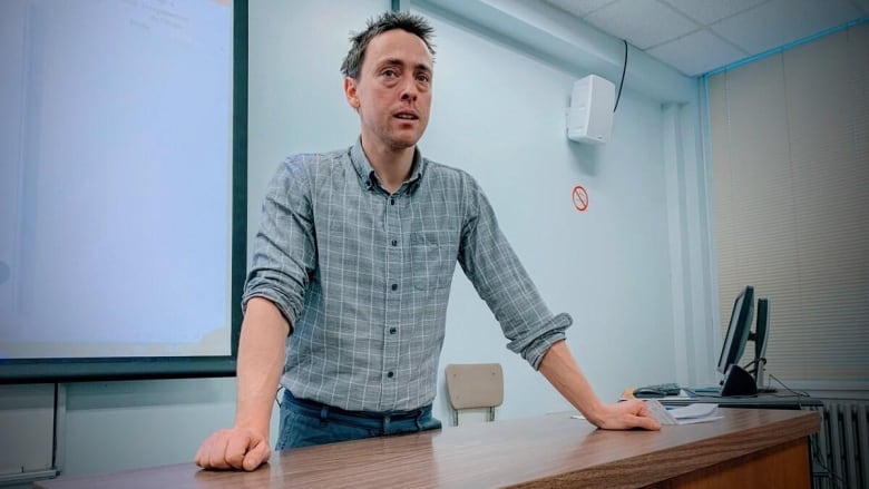 A man in a short stands behind a desk in a classroom.