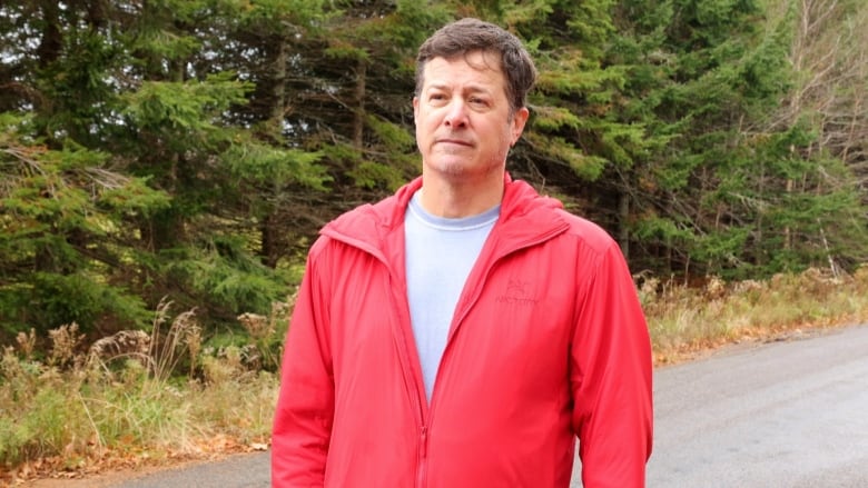 a man in a bright rain cost stands alonga  wooded  rural road. 