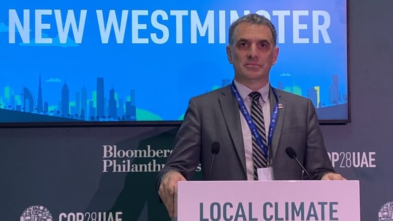 A man in a grey suit stands at a podium with a blue screen behind him. It displays the words New Westminster. Other signage says COP28 UAE.