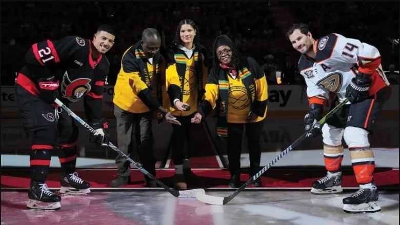 Three people dropping a puck for two fully-dressed hockey players.