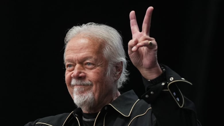 A man with gray hair and blue eyes holds up his hand with two fingers up making a peace sign