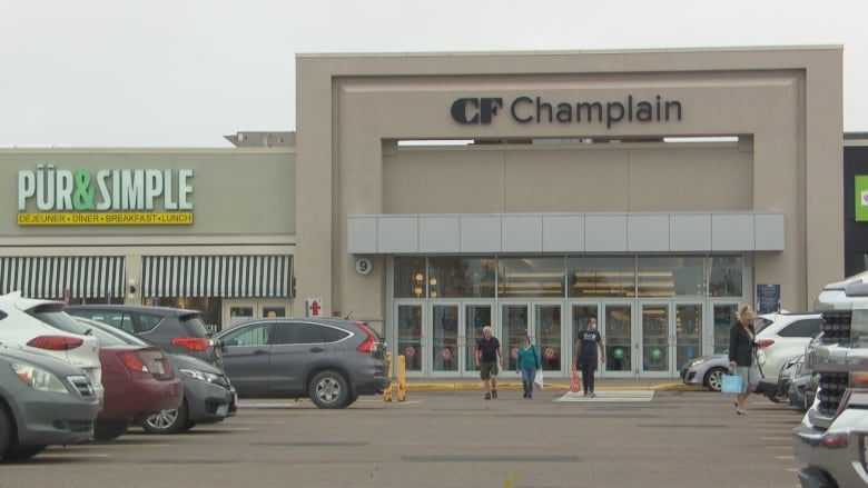 A large beige building with a sign that says Champlain Mall. People and cars are in front of it.