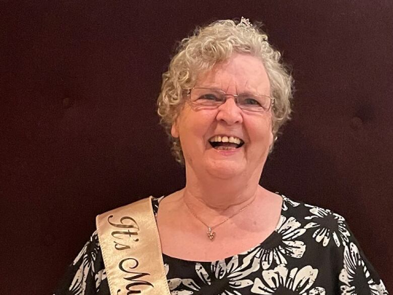 A senior woman with short, curly white hair and rectangular glasses smiles.