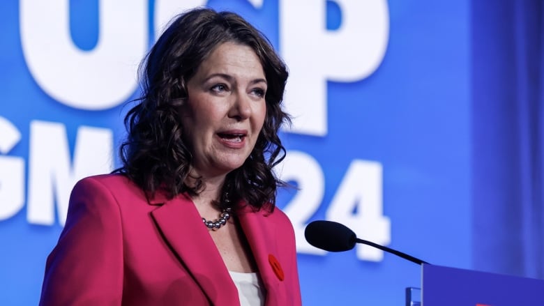 A woman, Danielle Smith, stands at a podium. There is a large blue background behind her that says 