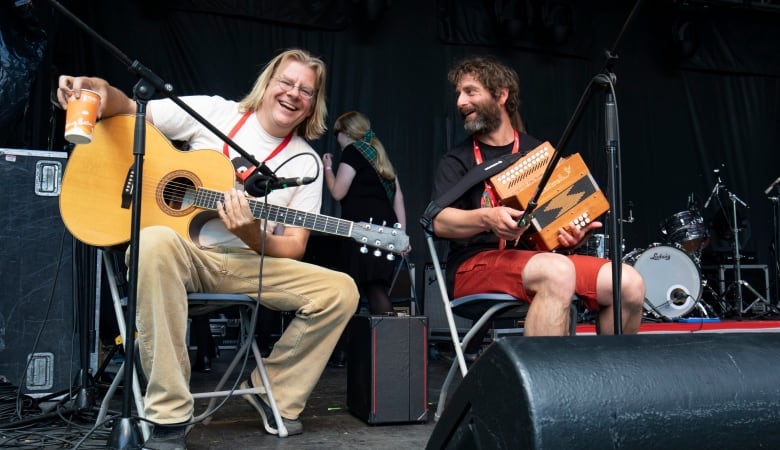 Two people sitting on chairs on a stage. One is holding a guitar and smiling with a coffee cup in his hand. The other is holding an accordion.