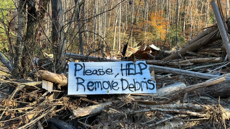 A sign reading, 'Please help remove debris,' is seen placed atop a pile of wood and branches.