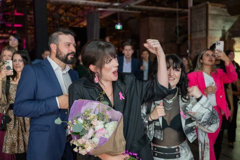 A woman, centre, in a crowd of people, cheering while holding a bouquet of flowers