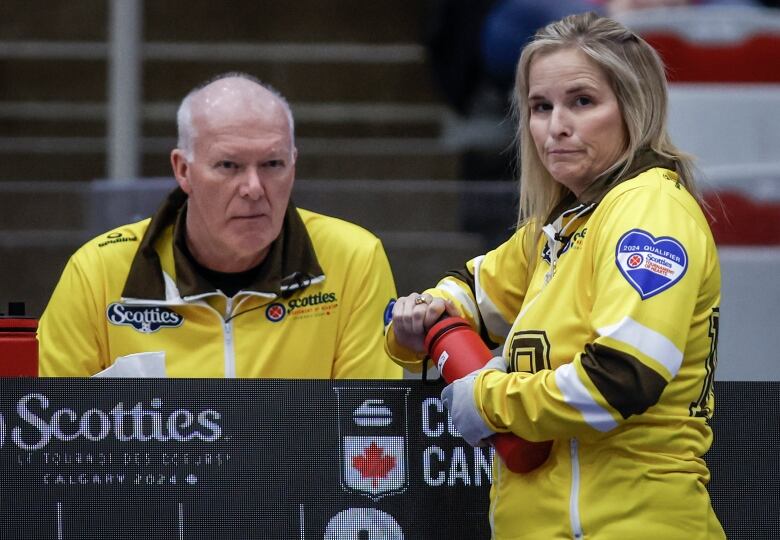 A man and woman stand next to each other wearing matching yellow jackets at a curling final. 