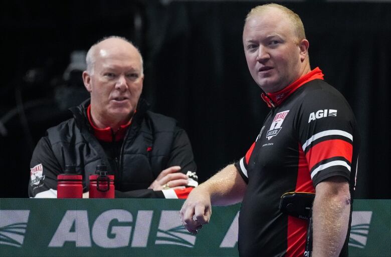 A father and son stand beside each other as they watch others curl during a sports tournament. 