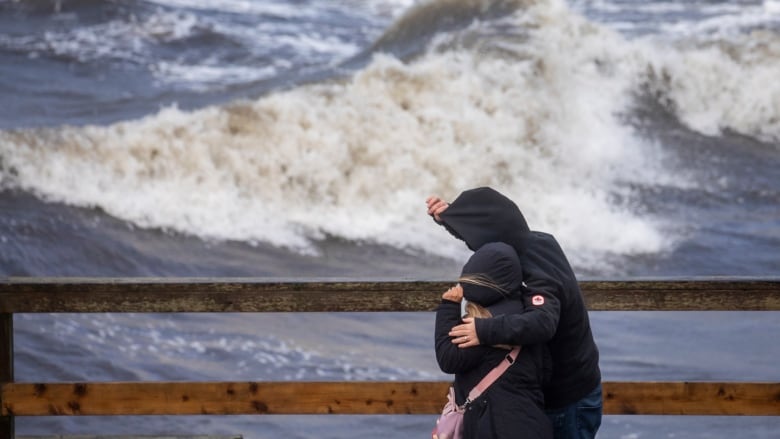 Two people cover their heads with their jacket hoods, while on a pier with a huge wave beside them.