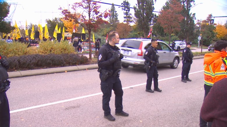 Officers stand on a road while protestors holding yellow flags gather behind them.