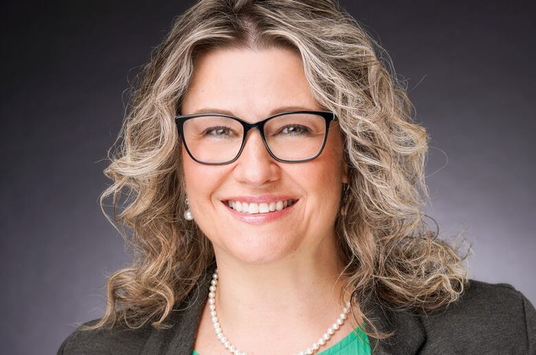 A smiling woman with wavy blonde hair wearing a glasses, a green shirt, and a brown blazer with pearls.