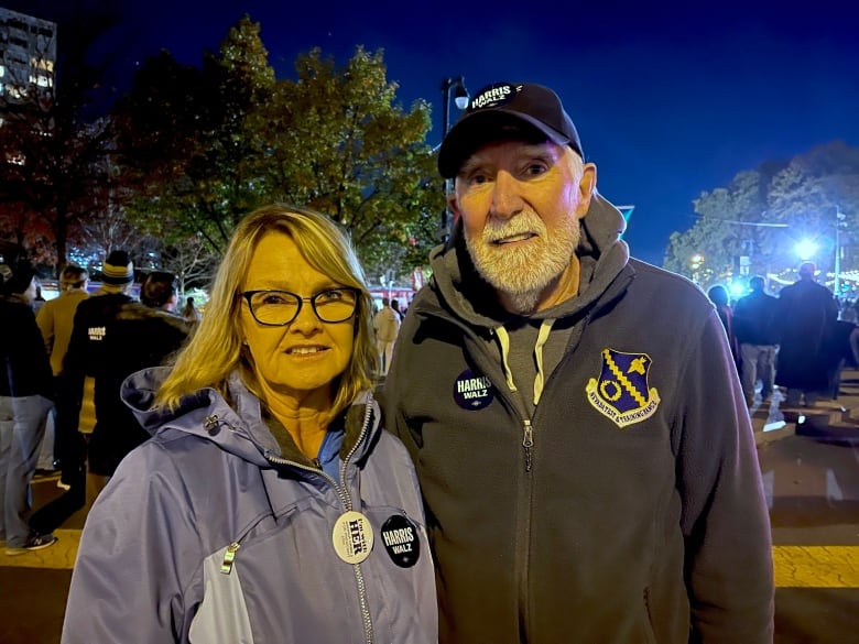 A man and a woman pose. They wear pins and a hat with Kamala Harris messaging.