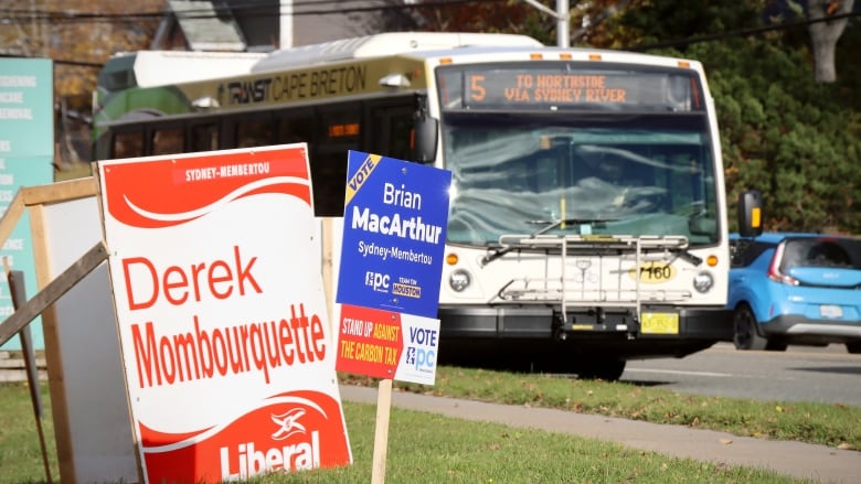 A large white sign with red lettering is on green grass next to a smaller blue sign with white lettering as a transit bus drives past.