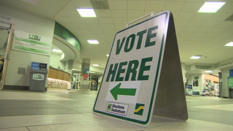 A white a-frame sign with green text sits in an open room. The text says VOTE HERE. A green arrow is below pointing left