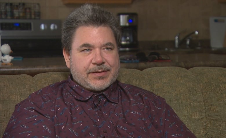 A man with short, spiked grey-black hair sits on a couch. He wears a burgundy button-up collared shirt.