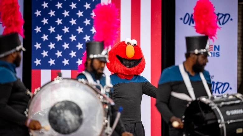 An elmo dances on stage with a marching band in front of an American flag