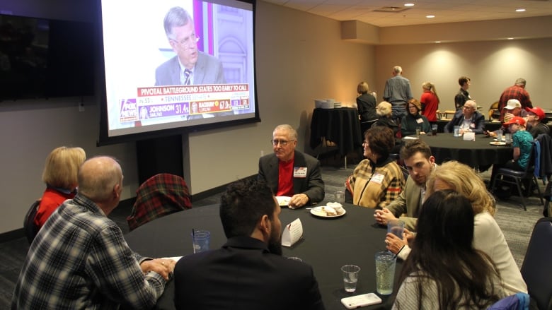 People sit around round tables and watch a projection screen showing an election night broadcast.