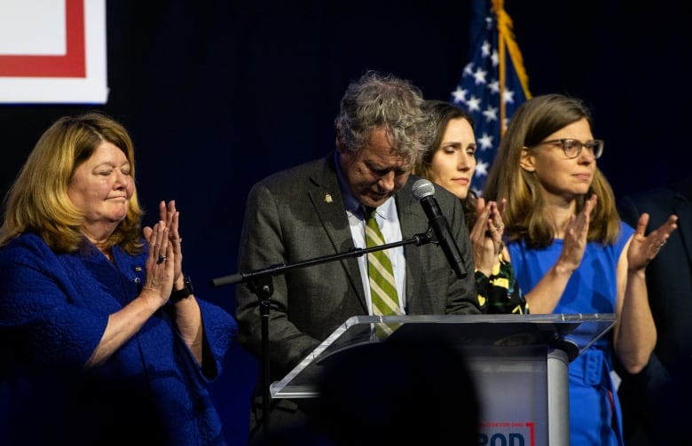 A man with grey-brown hair bows his head while standing at a podium as three women standing near him cap.