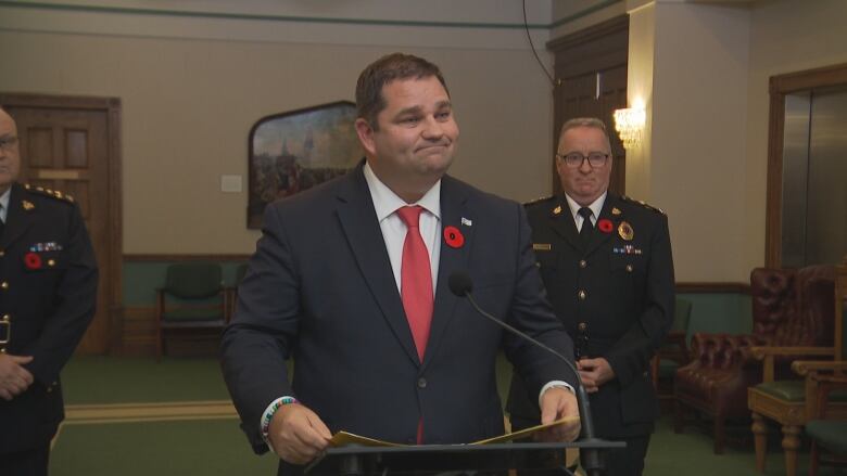 A smiling man wearing a suit stands in front of a lectern. Two police officers in uniform stand behind him.