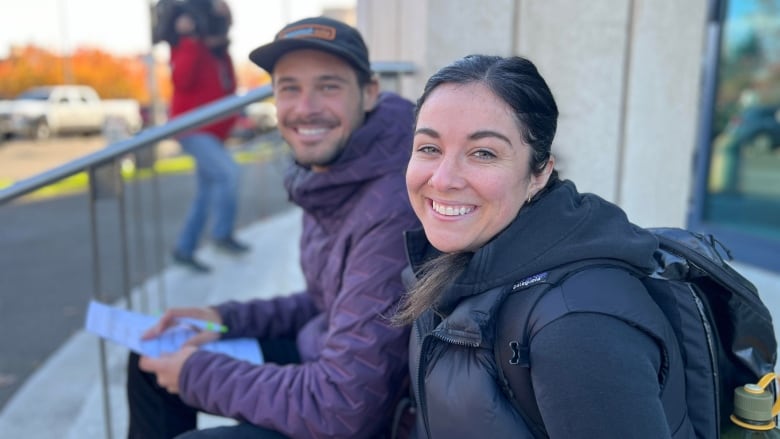 A man and a woman in winter clothing smile for the camera, sitting on steps outside a polling place. The man holds a sheet of paper in his hand.