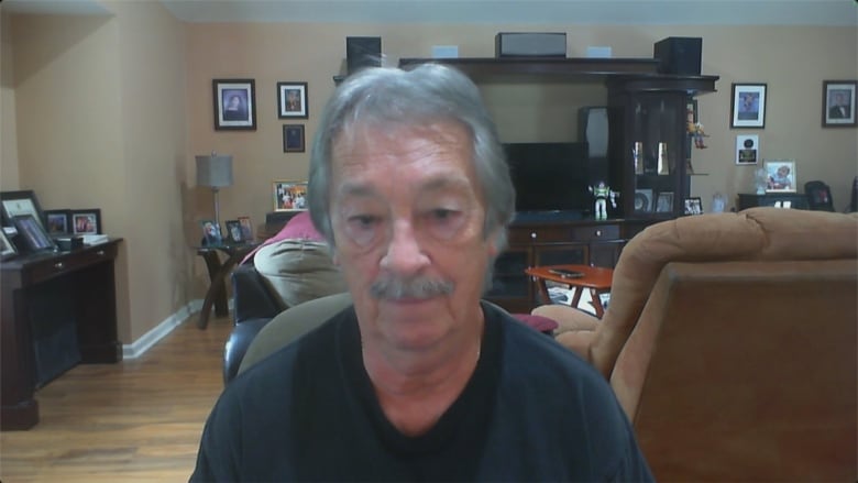 A man wearing a black t-shirt sits in his living room.