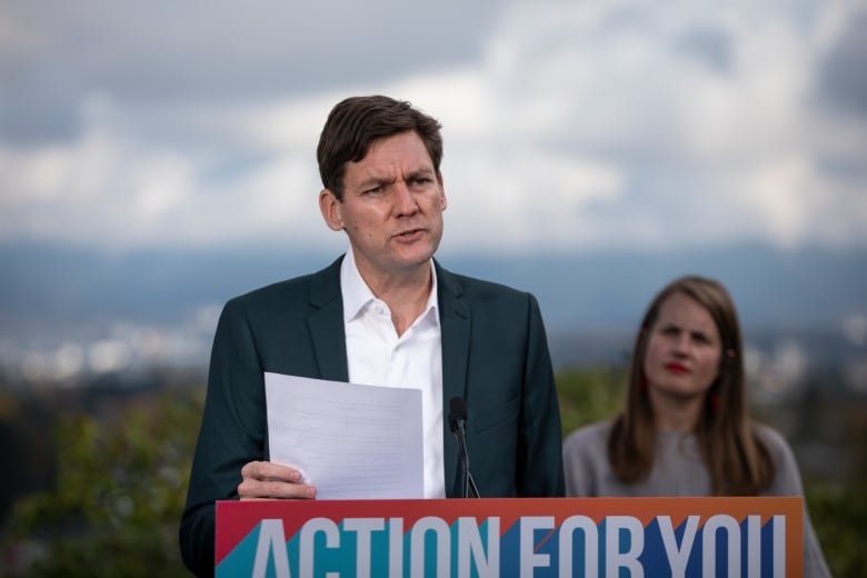 A man delivers a news conference at a podium marked 'Action For You', as a woman looks on.