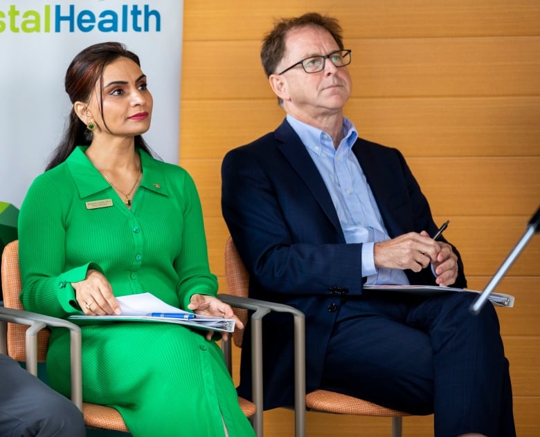 Two men and a woman sit on chairs in front of a banner reading 'Vancouver Coastal Health'.