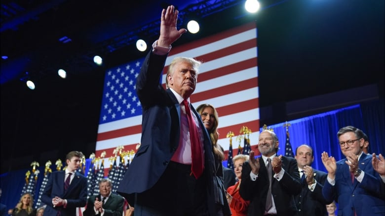 A man in a suit on a stage waves in front of a group of applauding people on the stage.