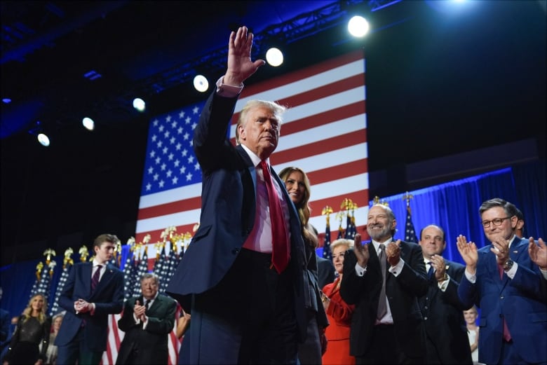 A man in a suit on a stage waves in front of a group of applauding people on the stage.