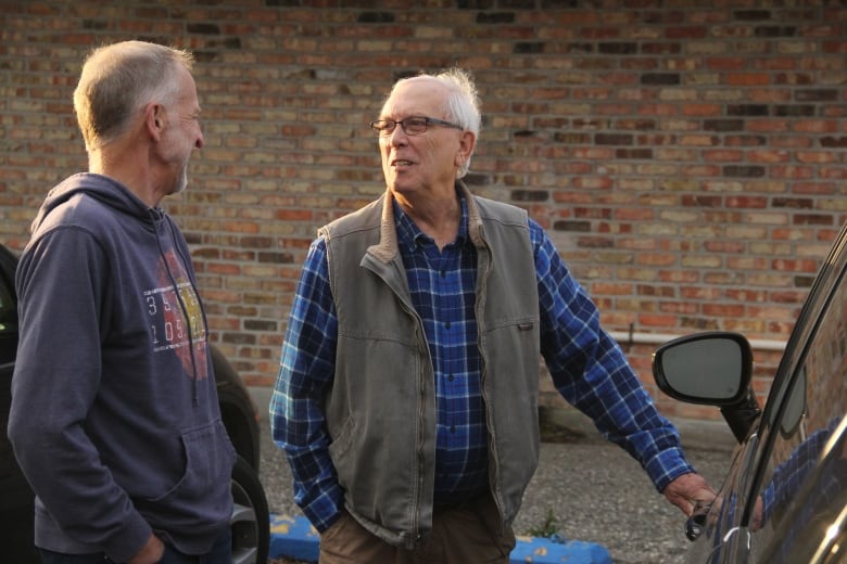 A man in a grey bomber jacket over his plaid blue shirt has his left hand holding the door, while speaking with a man to his right.