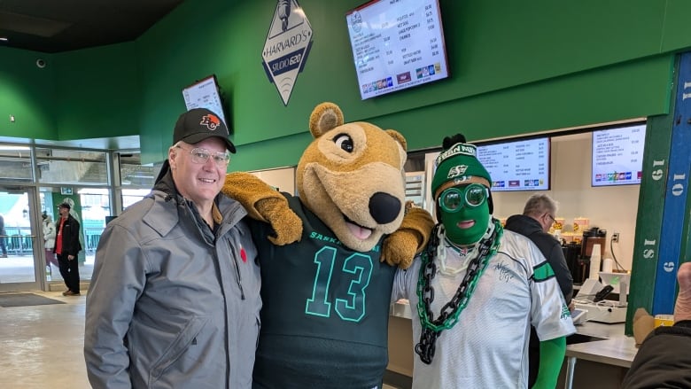 A white-haired man in a grey coat and black hat stands beside a man in a gopher mascot outfit and a man in a white jersey and green facepaint.