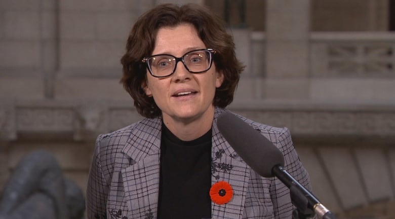 A woman with short, dark wavy hair and eyeglasses speaks from behind a podium.