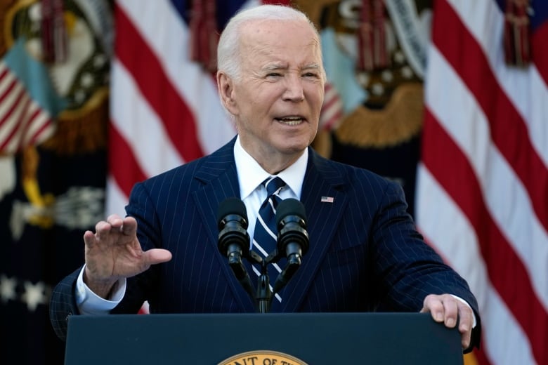 An older white haired man who is clean shaven and wearing a suit and tie speaks outdoors behind a podium.