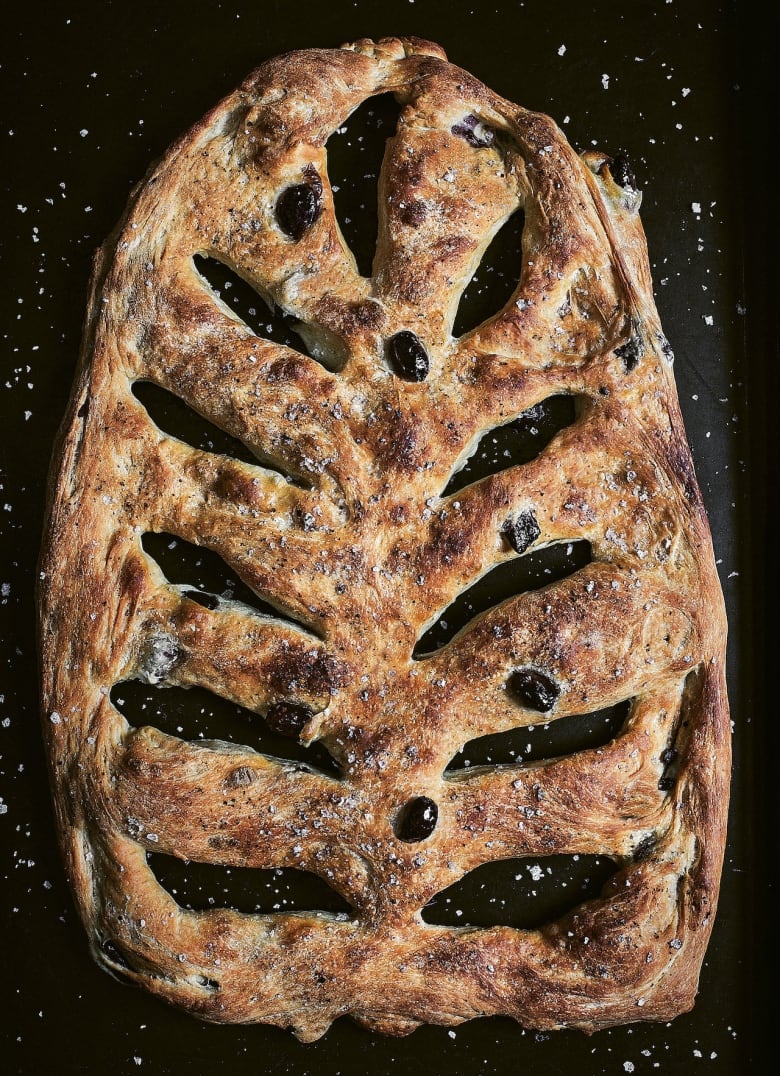 A flatbread topped with salt and black olives, with decorative openings cut into the dough on the diagonal. It's on a black background. 