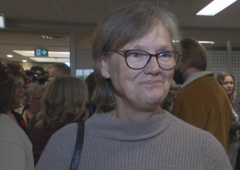 A woman with glasses in a brown sweater stands in a crowded room talking to a reporter.