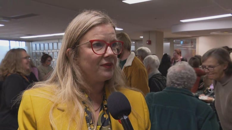 a woman with a bright yellow blazer and red eyeglasses stands in a crowded room speaking into a microphone.