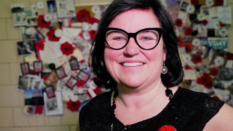 A smiling female teacher wearing a beaded poppy on her dress stands in a hallway, a bright mural of archival images, writing and poppies behind her.