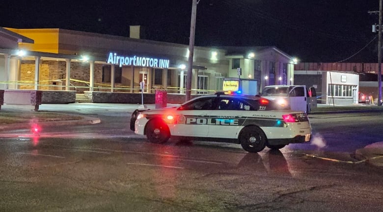 A police car is parked near a taped-off road. It is night time. The lights of a hotel can be seen in the background.