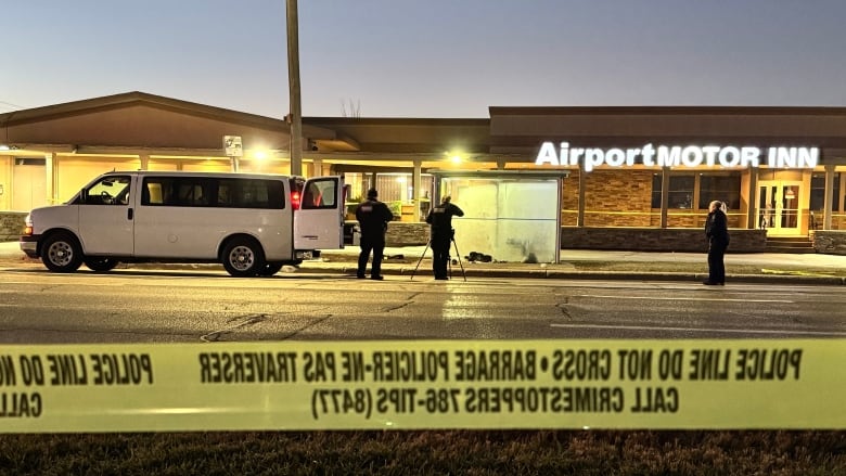 A band of yellow police tape is in the foreground.  Police officers in the background take photo at a frosted-up bus shelter in front of the Airport Motor Inn.