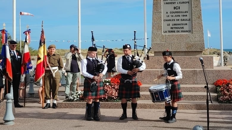 Three youths in kilts play bagpipes and drums in front of a memorial surrounded by flags.