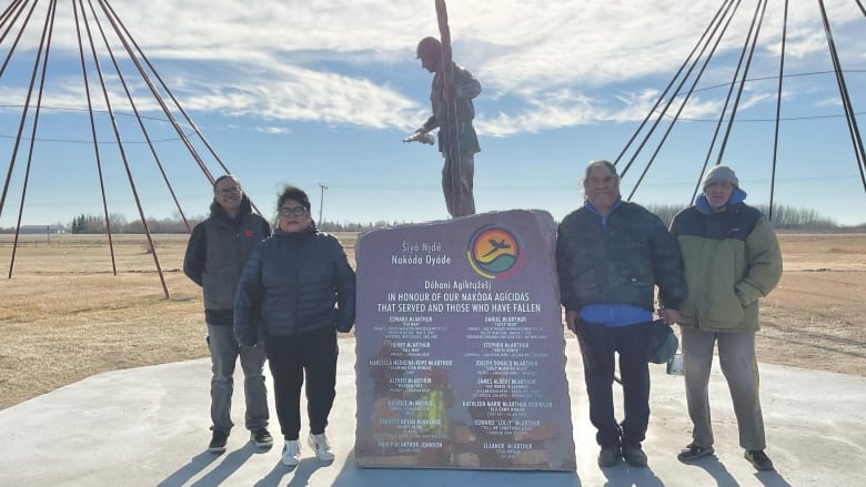 A woman with glasses and her hair up, stands around a stone and metal monument with three other men.