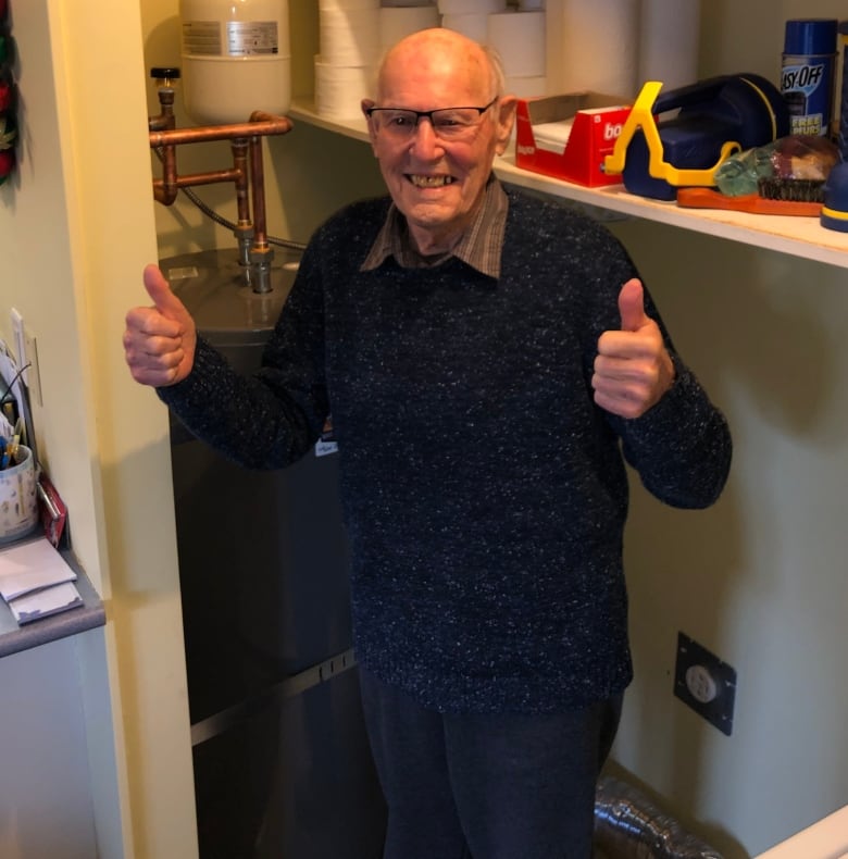 A senior man smiles and holds two thumbs up. Behind him is a special water tank gifted to him by a Victoria plumbing company. 