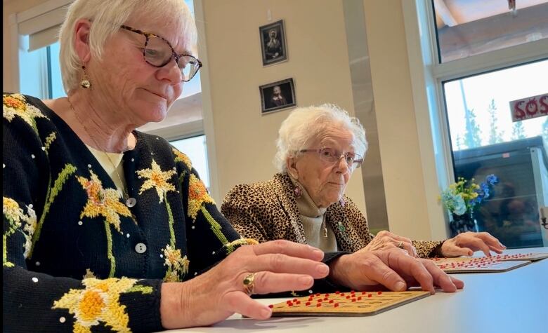 Two older women sit at a table playing bingo.