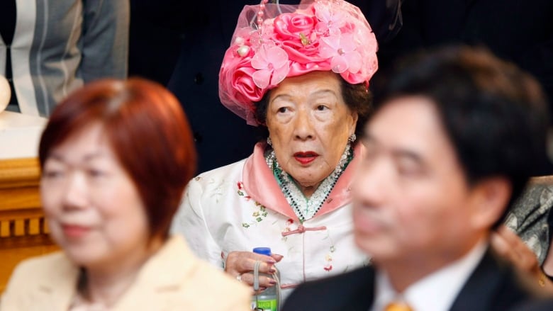 A woman in a large bright pink hat looks on from the back at a ceremony.