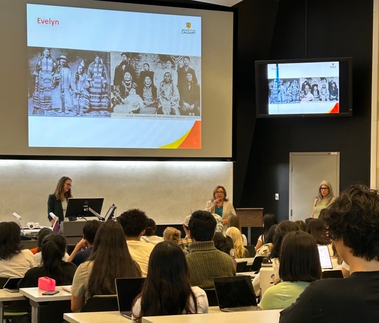 Three women stand at the front of a lecture theatre in front of a group of seated students.