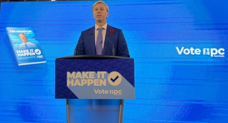 A man in a suit and tied stands behing a podium in front of a large blue screen flanked by Canadian and Nova Scotian flags.