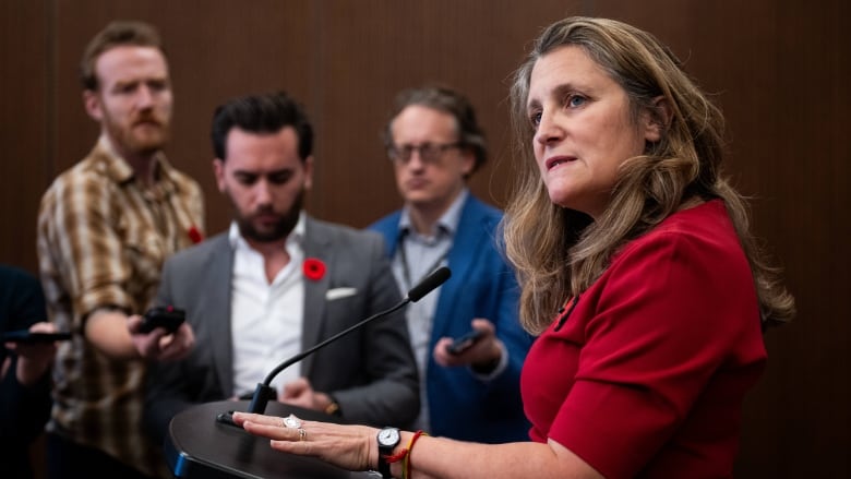 Chrystia Freeland, wearing a red shirt, speaks to reporters at a podium