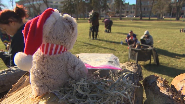 A teddy bear sits on a pile of wood, in front of it is a fire.